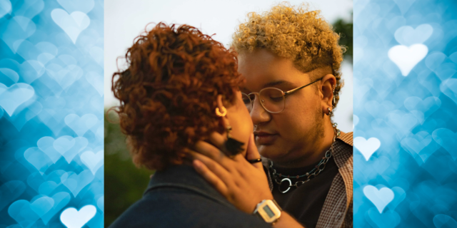 In the center of the image, there is a photo of a mixed race transmasculine person with a light beard and short, curly, blonde hair wearing a black T-shirt, an open, button-up tan shirt, glasses, a chain necklace, and a gold watch. They are standing outside and holding another person's face in their hands. The other person has short, curly, red hair and wears a black coat and dangling earrings. On either side of the photo, there are blue and white hearts.