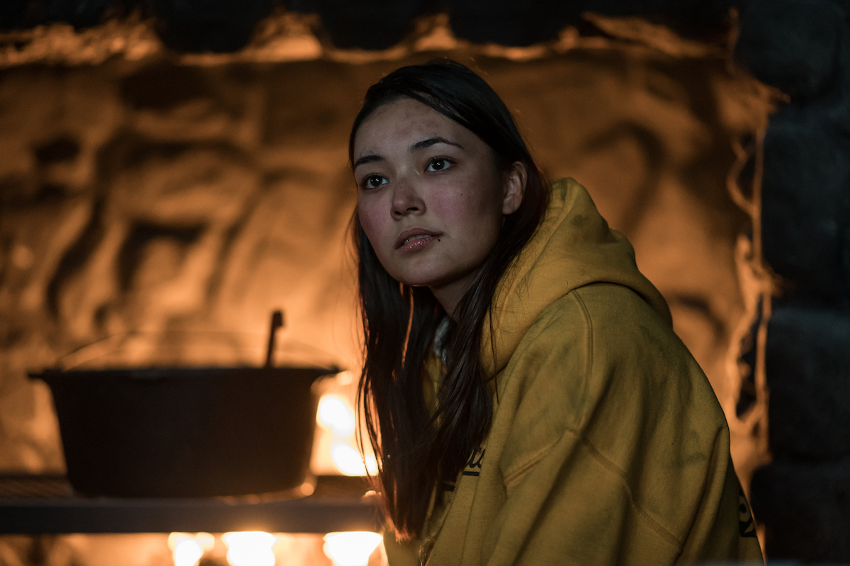 Alexa Barajas as Teen Mari in YELLOWJACKETS Season 2 wears a yellow hoodie near a fireplace. Photo Credit: Kailey Schwerman/SHOWTIME.