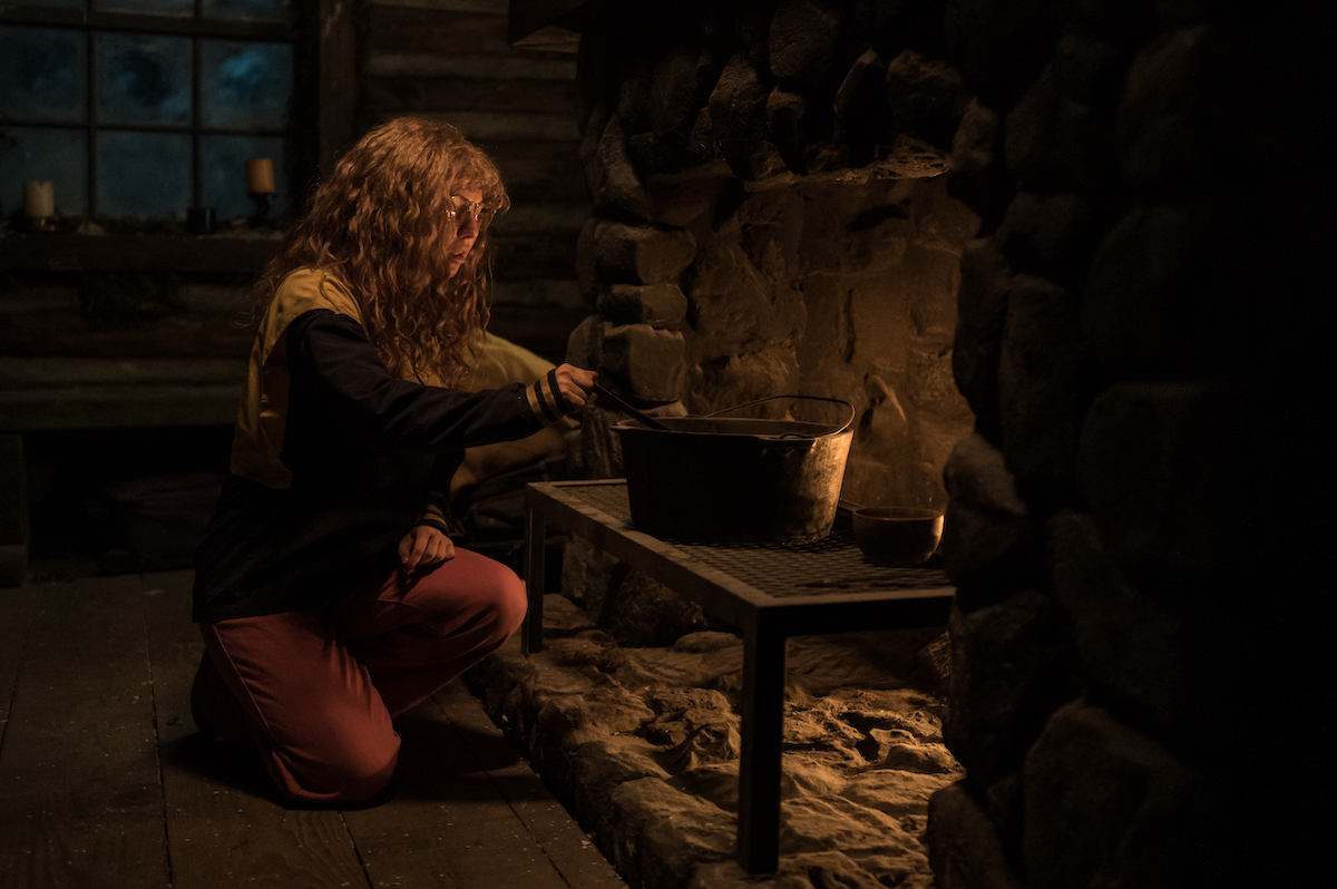 Samantha Hanratty as Teen Misty in YELLOWJACKETS Season 2 kneeling in front of a fire cooking in the cabin. Photo Credit: Kailey Schwerman/SHOWTIME.