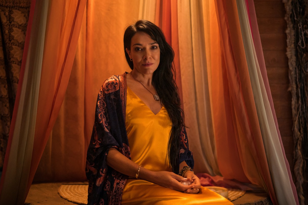 Simone Kessell as Lottie in YELLOWJACKETS Season 2 sits among colorful drapes. Photo Credit: Kailey Schwerman/SHOWTIME.
