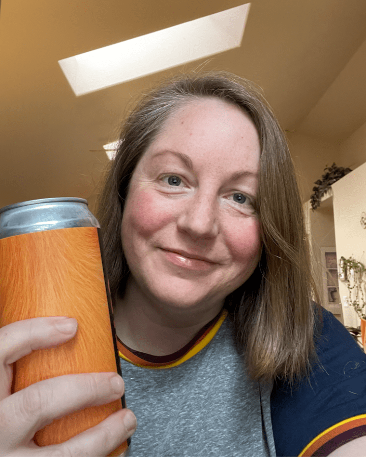darcy a white person with shoulder length light brown hair holds up a can of what looks like beer to the camera smiles lightly. they are wearing a ringer tee shirt where the rings are rainbow