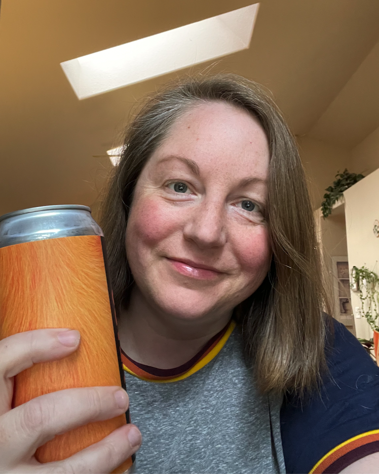darcy a white person with shoulder length light brown hair holds up a can of what looks like beer to the camera smiles lightly. they are wearing a ringer tee shirt where the rings are rainbow