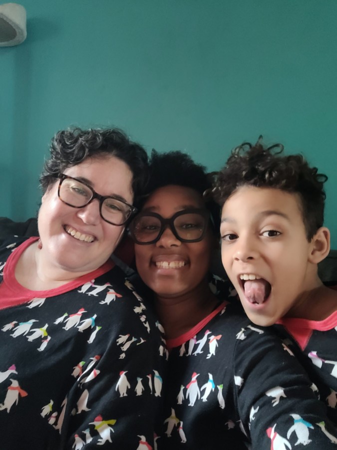 a turquoise background. three people in matching black pajamas with white penguins on them. one white woman with short brown hair and glasses smiling, one black woman with short brown hair and glasses smiling, one fair skinned boy with short brown hair sticking his tongue