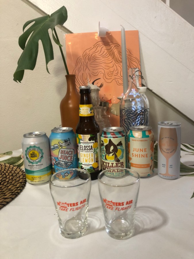 two cans of sparkling rose, two cans of hard kombucha, a can of hard seltzer, and a can of cream ale. there are two small Hooters glasses in front of the drinks.