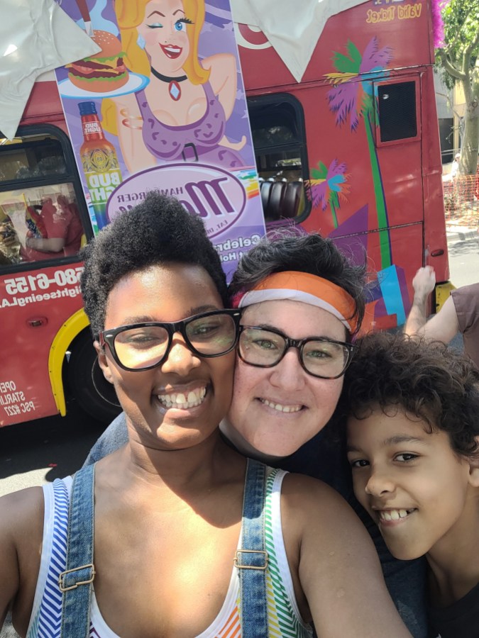 a red tour bus with a poster advertising Hamburger Mary's in pastel pinks and purples in the background. one black woman with short brown hair and glasses smiling. she is wearing a rainbow striped sports bra and overalls. a white woman with short brown hair and glasses smiling. she is wearing a lesbian flag bandana and a grey shirt. one fair skinned boy with short brown curls smiling in a black shirt