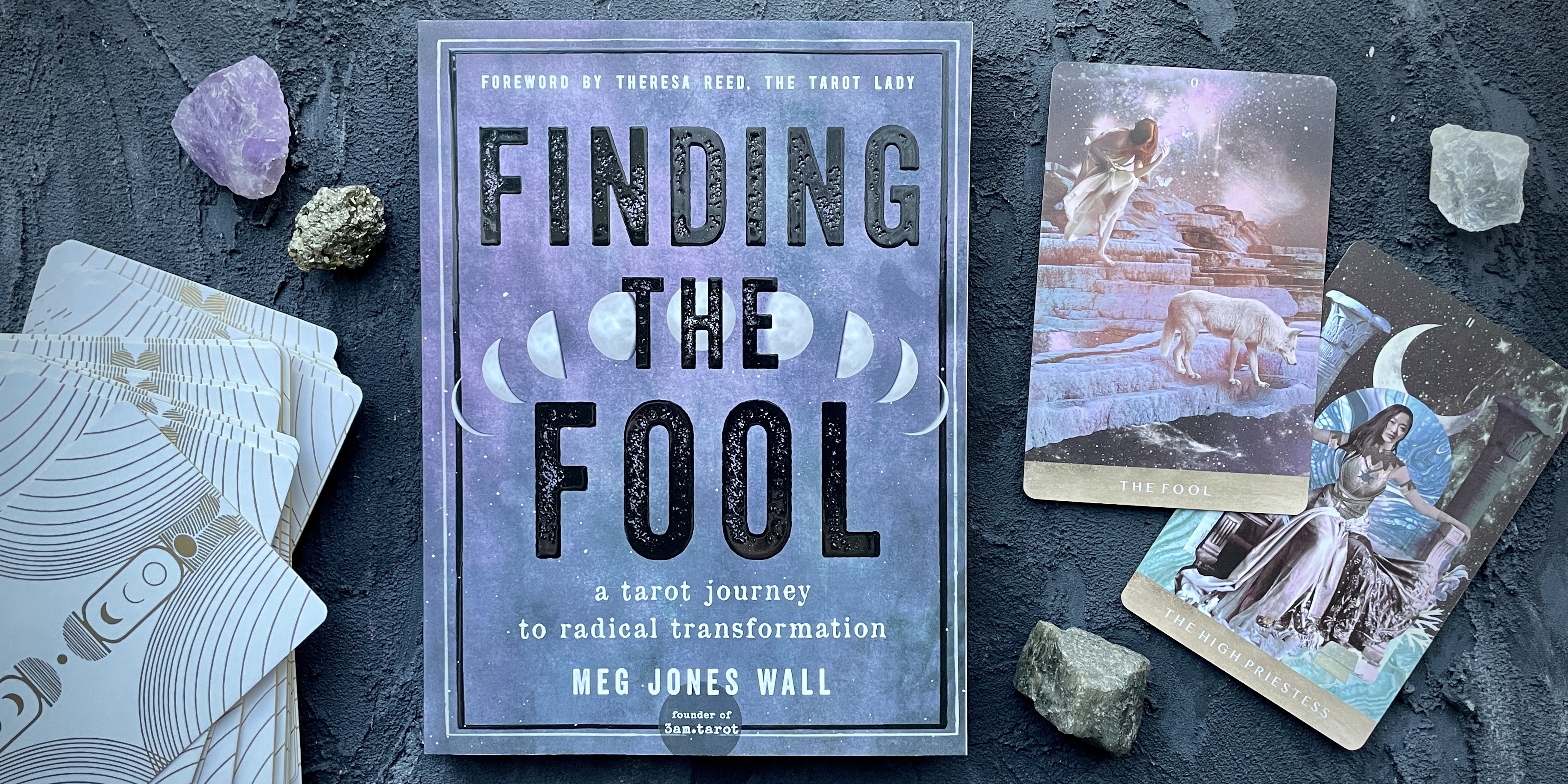 a gorgeous layout of the Finding the Fool book which is a gradient blue to purple with moon phases on it, set against a background of color coordinating tarot cards and crystals