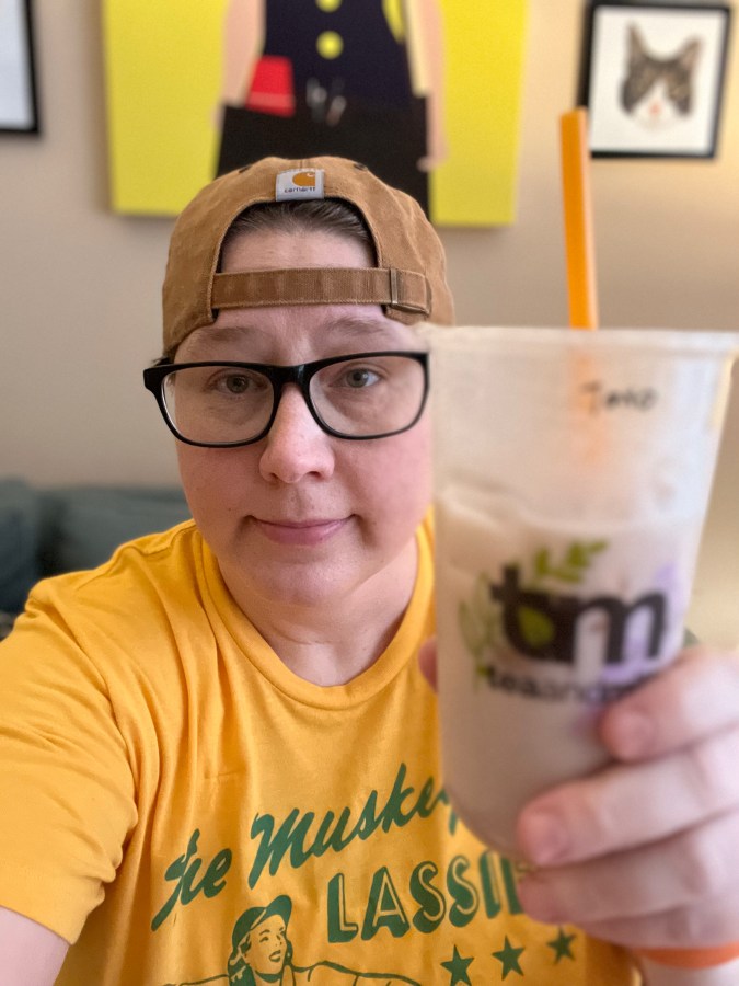 Heather a white butch lesbian holds up a packaged beverage that could be a bubble tea to the camera and smiles. she is wearing a backwards baseball cap and glasses
