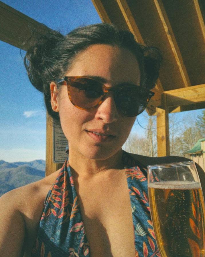 Kayla is a South Asian woman who here has her long dark hair up in two buns and is wearing sunglasses and a patterned bathing suit and holding up a glass of sparkling wine to the camera.