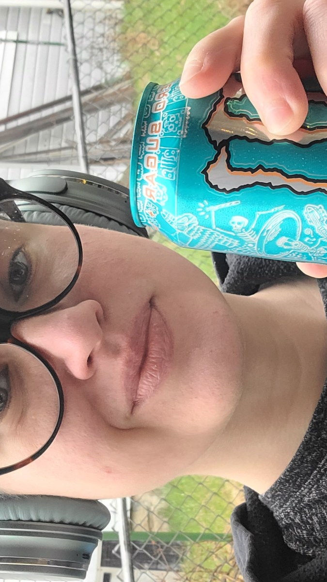 Nico is a white genderqueer human who is here wearing a trucker hat, headphones, glasses and a black sweatshirt and is holding up a monster can to the camera. they have shaved sides and wavy brown hair on top.
