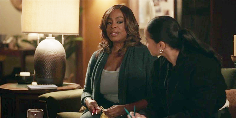 The Rookie: Feds - Simone raises a skeptical eyebrow as Naomi sits down next to her and tries to intoce her to the fine art of knitting. Simone is sitting on the left side of the couch, wearing a soft tee and cotton cardigan while Naomi wears a black jacket, a barely visible white shirt and black pants.