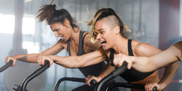 two women on exercise bikes screaming. one has a ponytail and one has an undercut