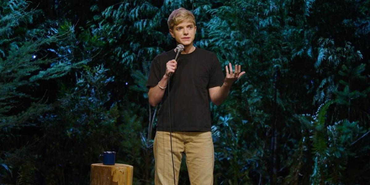 Mae Martin is on a stage with a mic, surrounded by trees