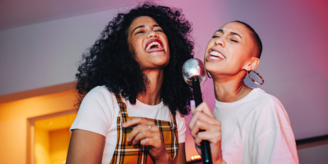a woman with curl hair and bright red lipstick and a plaid dress over a white tee sings karaoke with a woman with a shaved head in a white tee who is holding the mic
