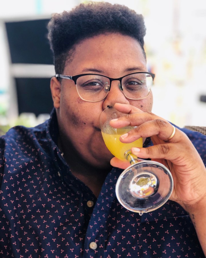 shea, a Black nonbinary human with short hair and glasses wearing a patterned button up smiles and drinks a mimosa 