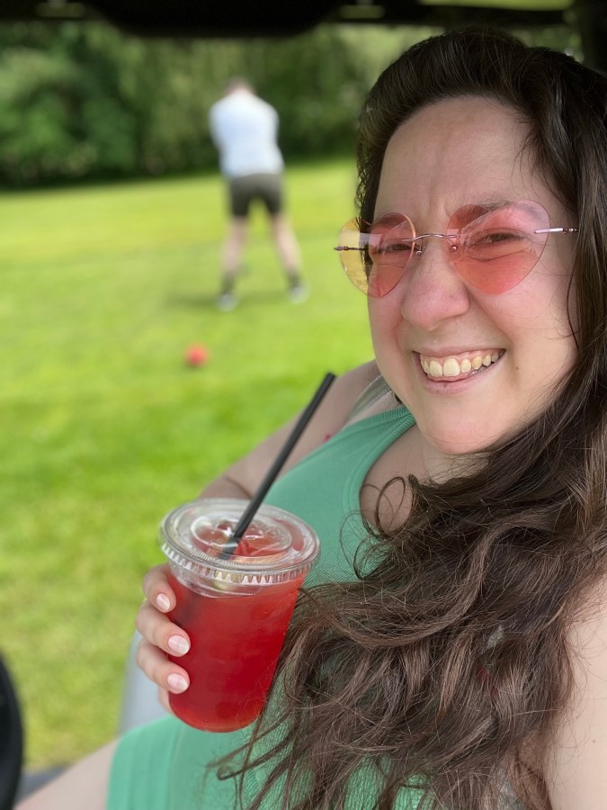 Vanessa, a white woman with long hair and heart shaped sunglasses, is smiling and holding a red beverage on what appears to be a golf course
