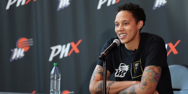 Brittney Griner in a Phoenix Mercury press conference, sitting at a podium table in a black t-shirt, smiling with her arms crossed.