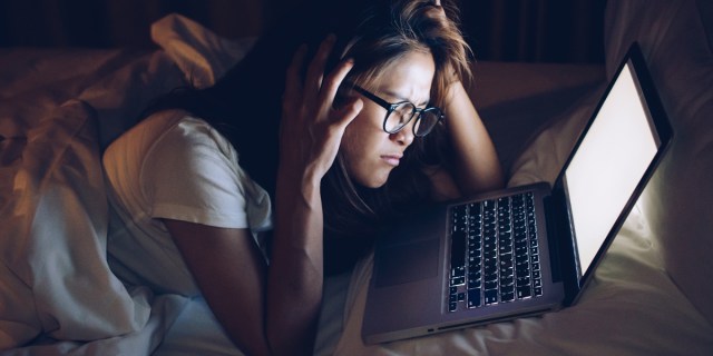 An Asian woman with glasses cannot sleep and is frustrated looking at a computer instead.