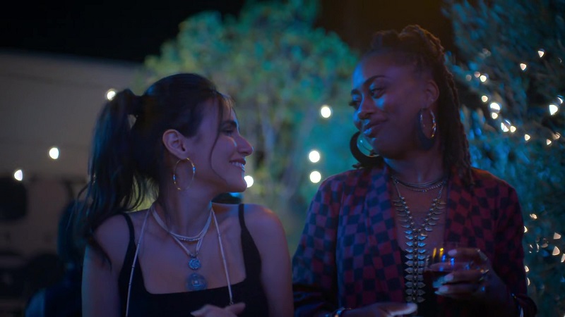Angelica slides in next to Malika and agrees to be her wing-woman at Malika's first queer party as a baby gay. Angelica is on the left, smiling brightly while looking at her ex. Malika smiles back, while holding a glass of wine in her left hand and her phone in her right.