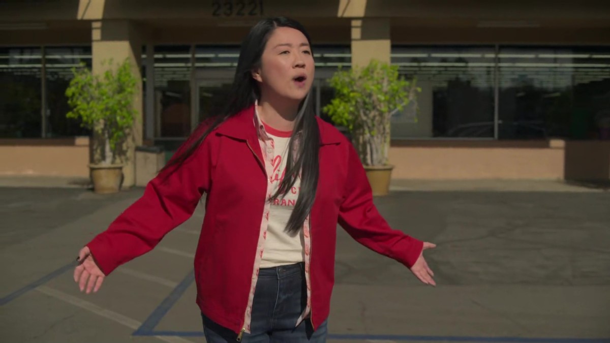 Alice stands in front of the grocery store, exasperated that her colleagues won't return to work. She's wearing a red and white ringer t-shirt, a red and white tartan button up and a red denim jacket. Her arms are raised in disbelief.