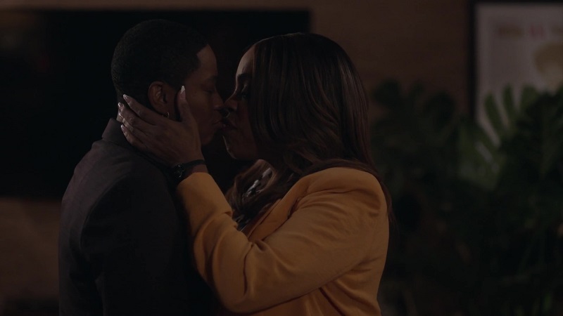 Simone pulls Dina into a kiss after returning from a stressful day at work. Simone is on the left, wearing a canary yellow blazer, while Dina is in all black on the left. 