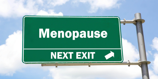 Against a blue sky filled with fluffy clouds, there is a green street sign with white text that reads, "Menopause: next exit." A white arrow points to the right.