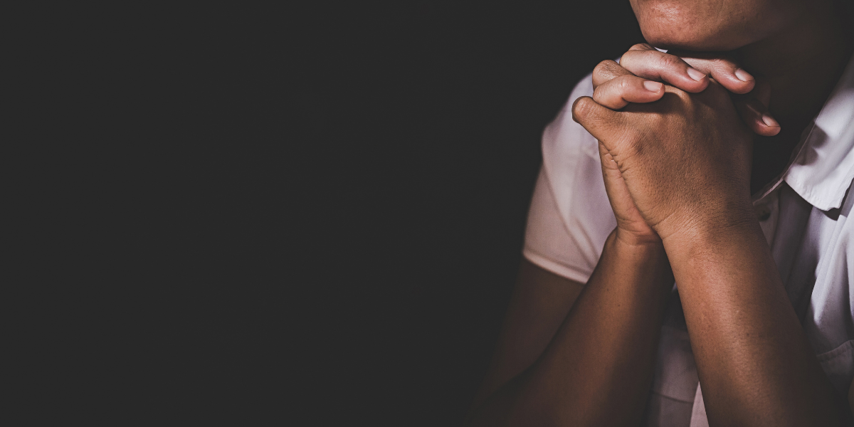The background is black. On the far right, there is the chin and upper toro of a Black woman wearing a short-sleeved, white collared shirt. She clasps her hands in prayer.