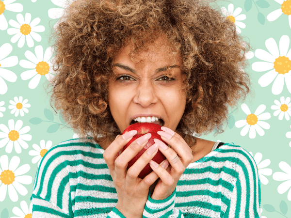 Against a light green background covered in drawings of daisies, a light-skinned Black woman with light brown, curly hair wears a green and white striped shirt. She bites into an apple.