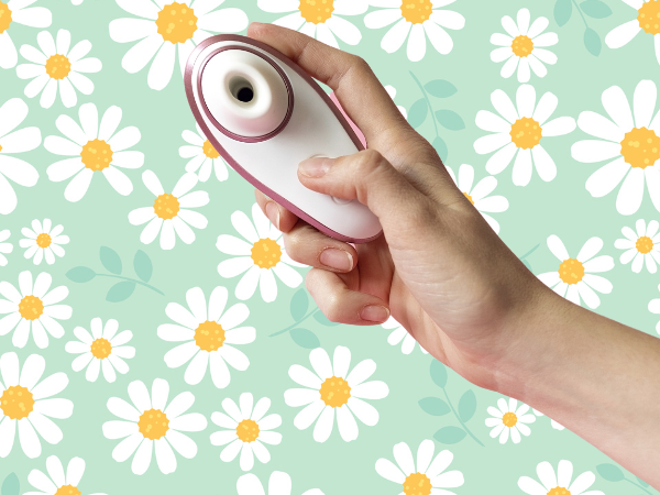 Against a light green background covered in drawings of daisies, a white woman's hand holds up a white and metallic pink suction sex toy, which can be used for clitoral or nipple stimulation.