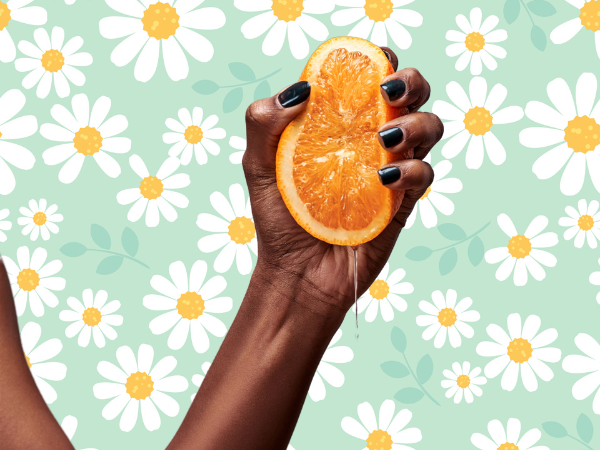 Against a light green background covered in drawings of daisies, a dark-skinned Black woman's hand with black nail polish squeezes half of an orange.