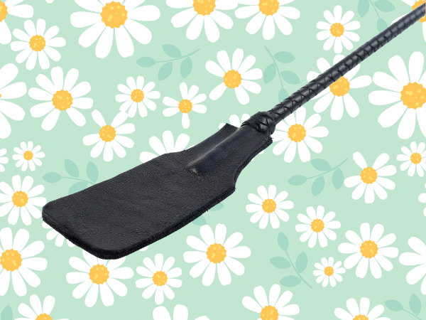 Against a light green background covered in drawings of daisies, there is the slapping portion of a black, leather riding crop and part of its leather handle.