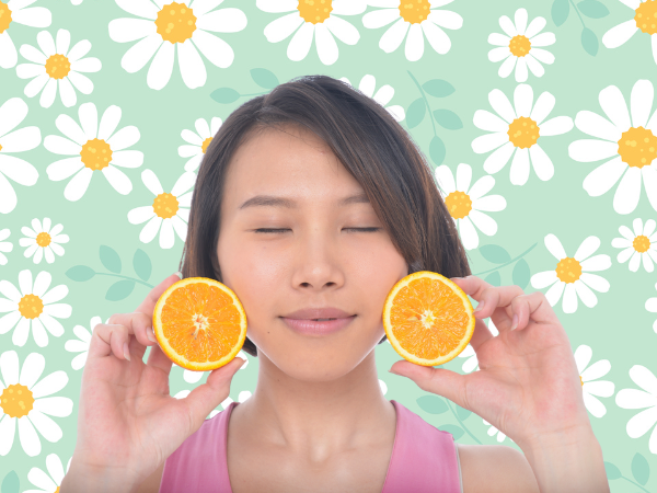 Against a light green background covered in drawings of daisies, an east Asian woman with a brown bob that's parted on the side wears a pink tank top. She holds up two halves of a clementine and closes her eyes.