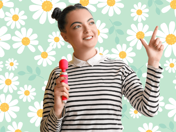 Against a light green background covered in drawings of daisies, an east Asian woman with green and black hair in two buns wears a black and white striped shirt with a white collar. She holds points up with her left hand. In her right hand, she holds a pink and white wand vibrator.