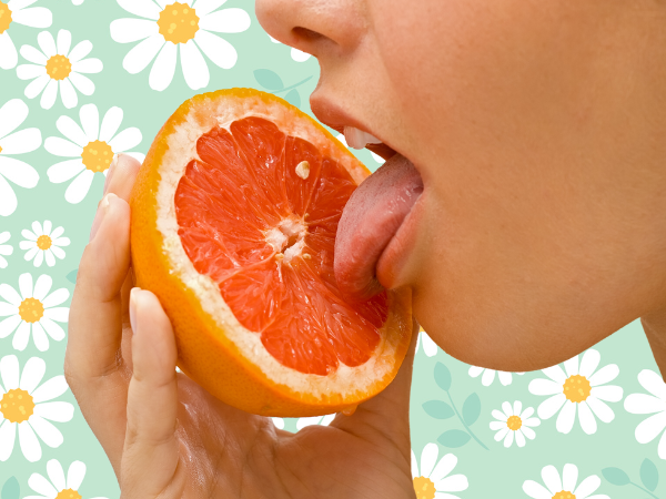 Against a light green background covered in drawings of daisies, there is the bottom half of a white woman's face. She holds up half of a grapefruit and licks it.