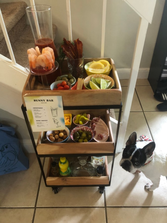 a rolling cart with ingredients for bloody marys on it and a little white and black french bulldog looking at it.