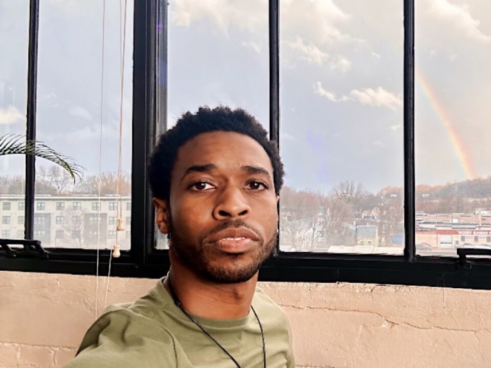 a portrait of Dior J. Stephens, a medium dark skinned black person with a small afro and a goatee, looking intently at the viewer. They are wearing a green shirt with a black necklace and a cloudy sky can be seen in a window behind them. 