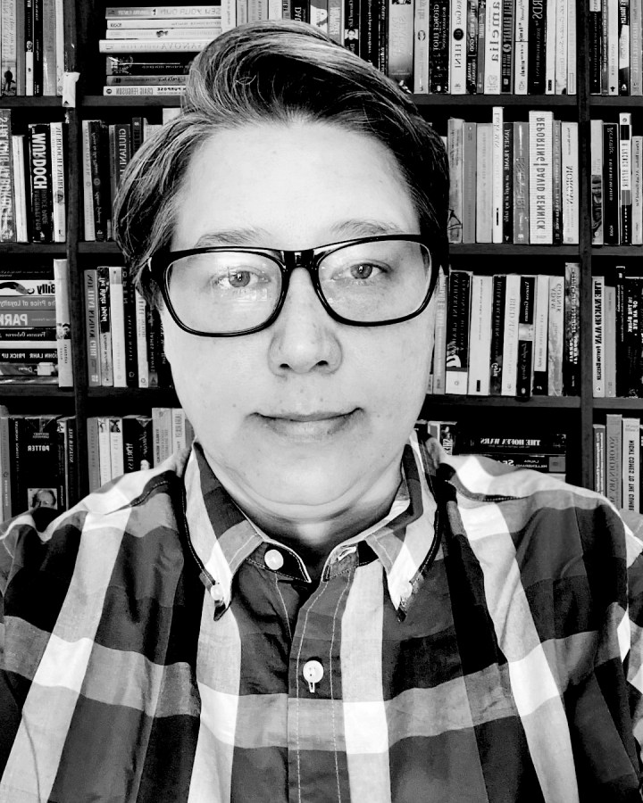 a black and white photo of Heather a soft butch woman with glasses wearing a plaid button up shirt and looking serious