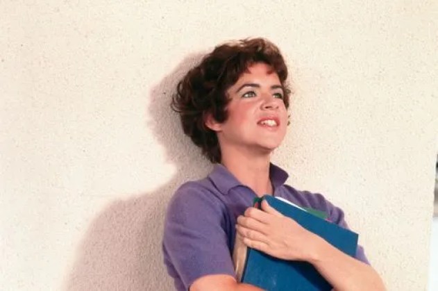 Rizzo in a purple polo shirt leaning against the wall in Grease