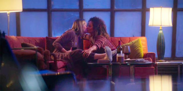 Stef and Lena kiss on the couch in front of big windows and a bottle of wine on Good Trouble