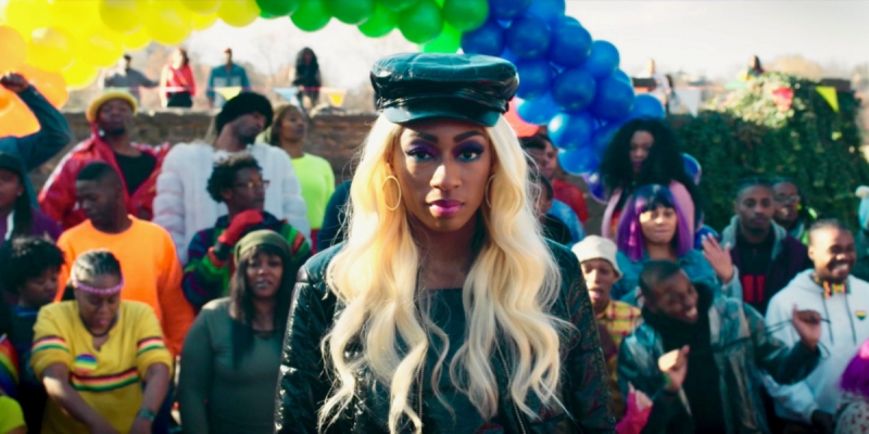 Tia from Boomerang, a Black lesbian with a long blonde wig, stands underneath a rainbow balloon arc during a Pride festival.