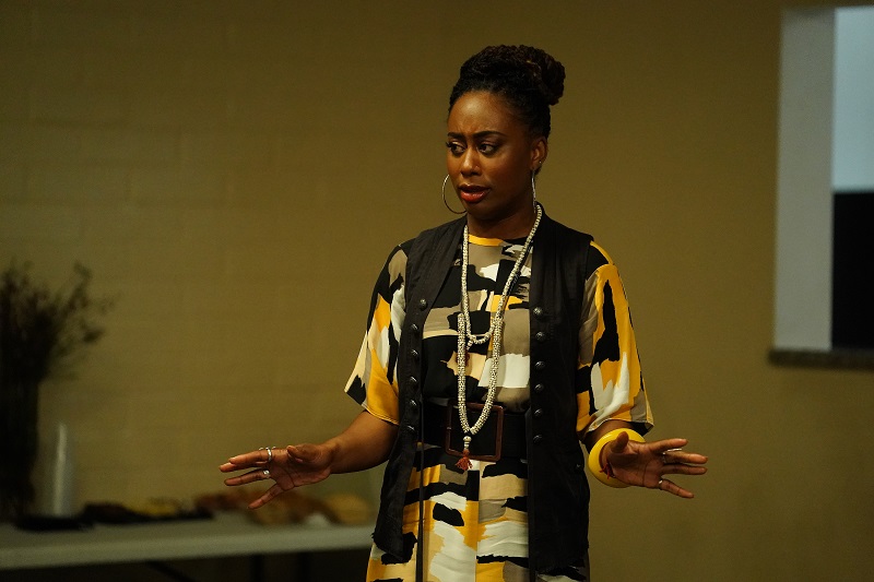 Malika hears pushback from the community where she's planning to locate her women's center. She's standing in a meeting room wearing a print dress with black vest and chunky black belt.