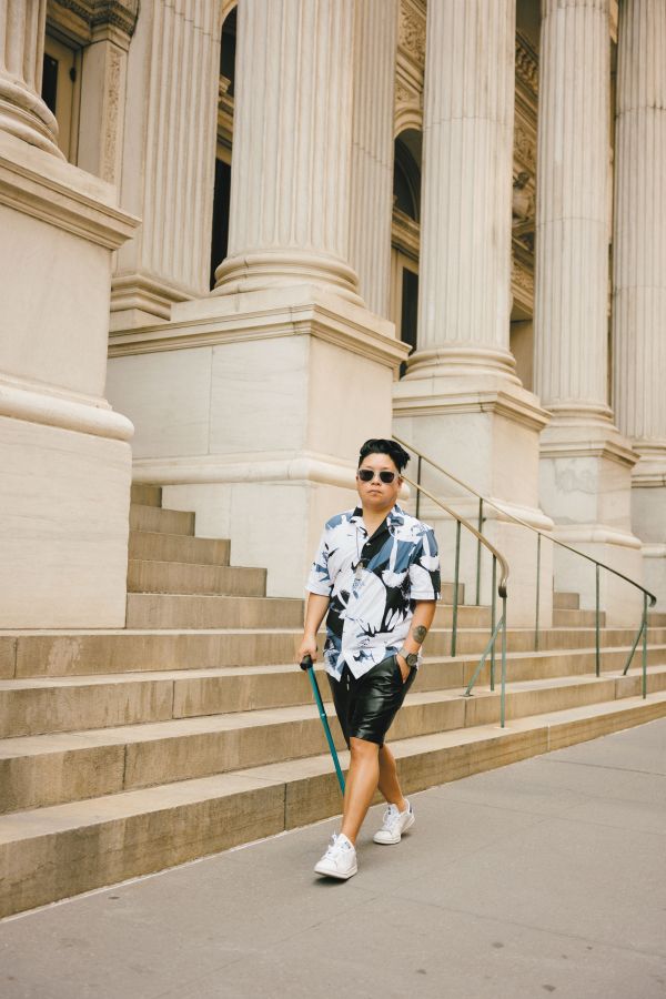 A brown trans person in black shorts, a large print button down, sunglasses, and white sneakers, using a blue cane, walks purposefully down a New York City sidewalk. They have one hand in their pocket, are looking directly at the camera, and seem confident and at ease.