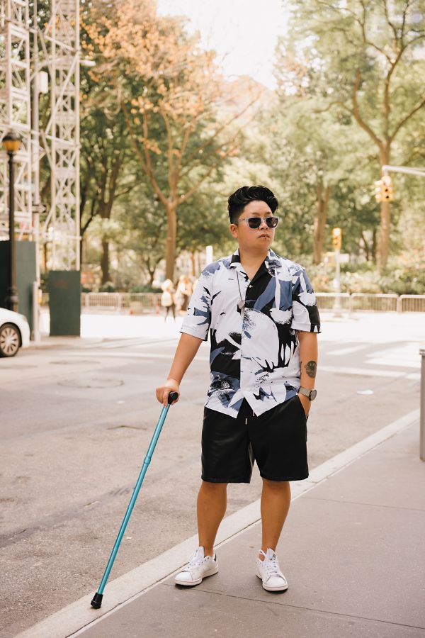 A brown trans person in black shorts, a large print button down, sunglasses, and white sneakers, poses on a New York City sidewalk. They hold their blue cane and look into the distance.