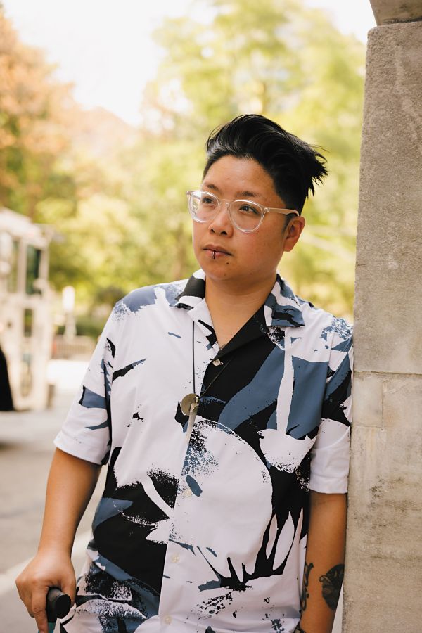 A brown trans person with a centered lip ring, wearing a large print button down and clear framed glasses, poses on a New York City sidewalk. They hold their blue cane and look into the distance.