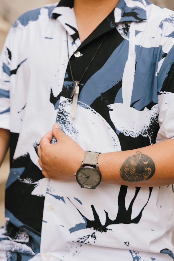A close up detail shot of a brown trans person wearing a blue, white, and black large print button down. We can see the details of their crystal necklace and their wrist watch.