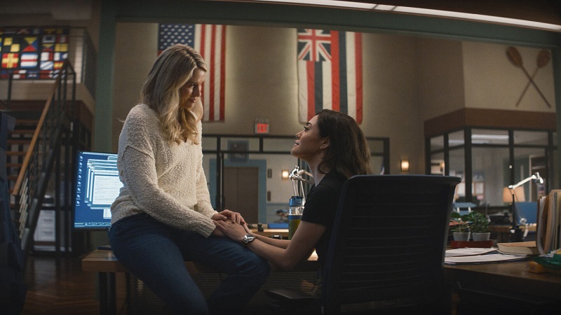 Kate sits on the edge of Lucy's desk, while Lucy is seated in her chair. They share a warm moment, celebrating their anniversary, while Lucy is on night watch duty.