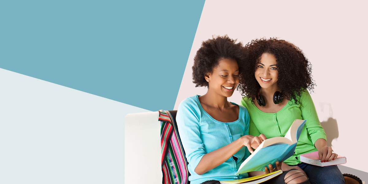 two Black women huddled close together reading a book together and smiling