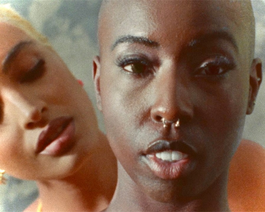 Two Black queer people holding each other while looking at the camera.