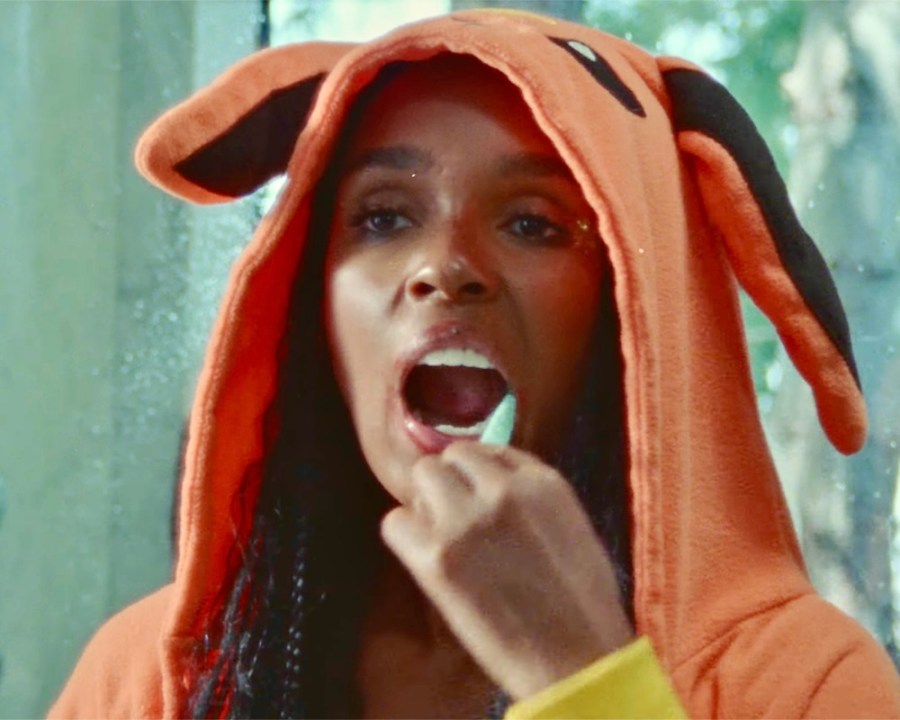 Janelle Monáe in a poker furry creature onsie, brushing their teeth. 