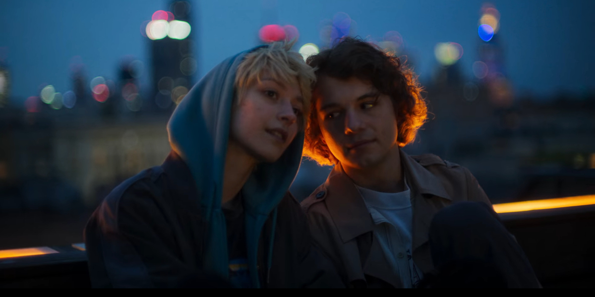 Alin Szewczyk as Tosiek and Jan Cieciara as Leon, lean their heads against each other at night with a city skyline behind them.