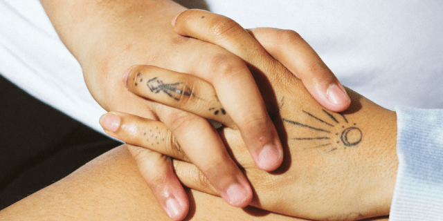 A close up image shows two light brown hands clasped together. One person wears a white shirt has short, unpolished nails. The other person wears a light blue sweatshirt and has slightly longer unpolished nails and black line tattoos on their fingers and palm.